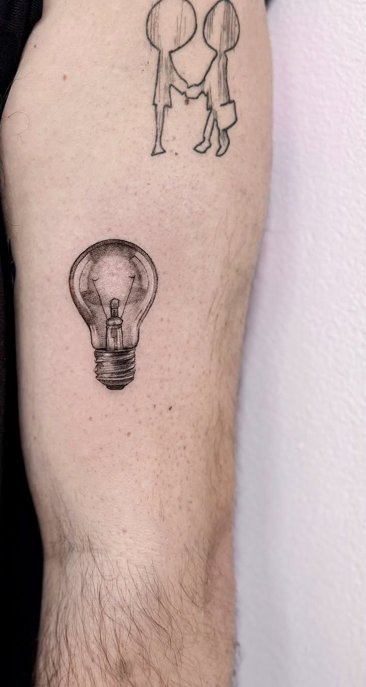 Lightbulb tattoo on a forearm with a small figure drawing above it.