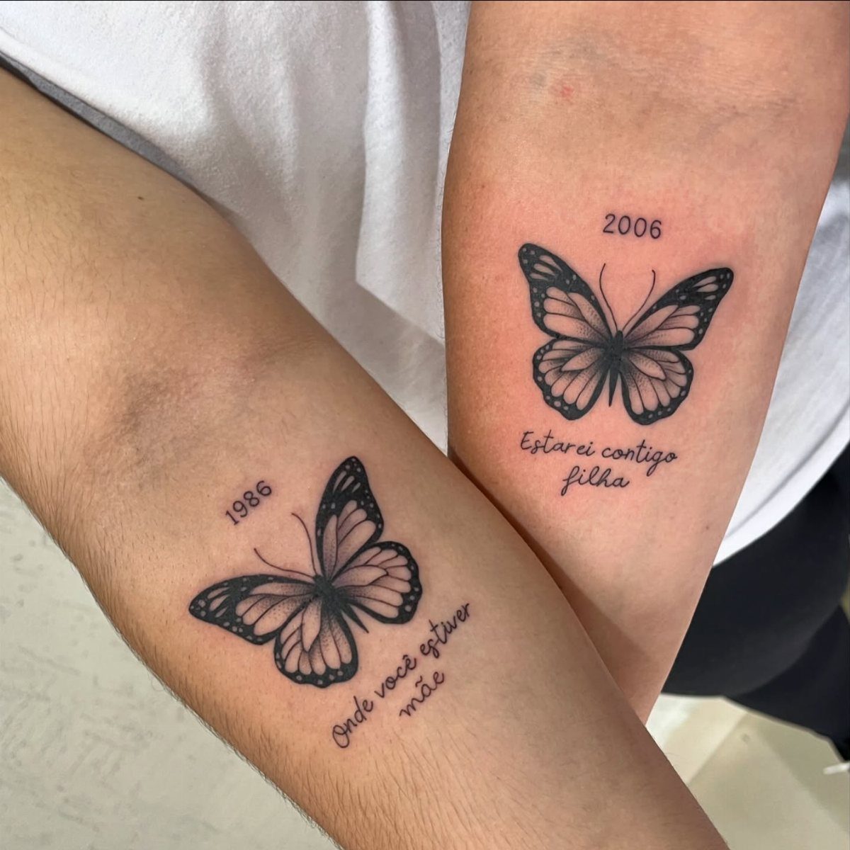 Two forearms with matching black butterfly tattoos and Portuguese text and dates.