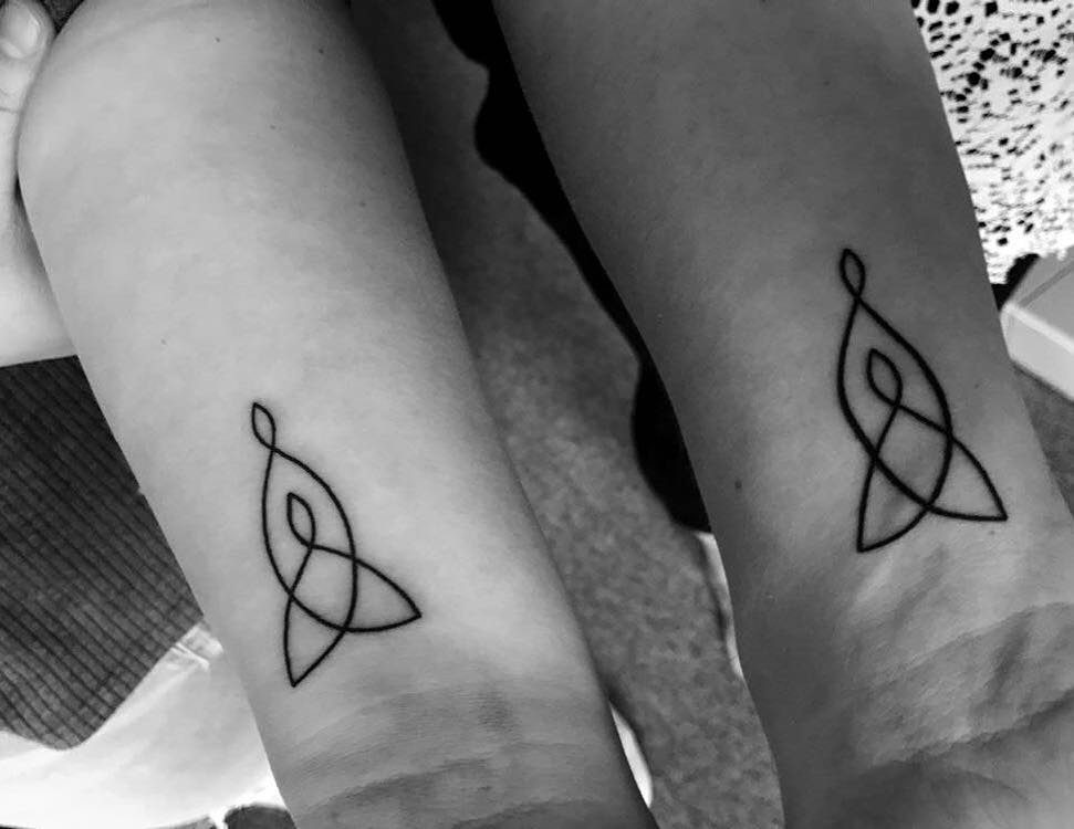 Two wrists with matching black ink Celtic knot tattoos in teardrop shapes.