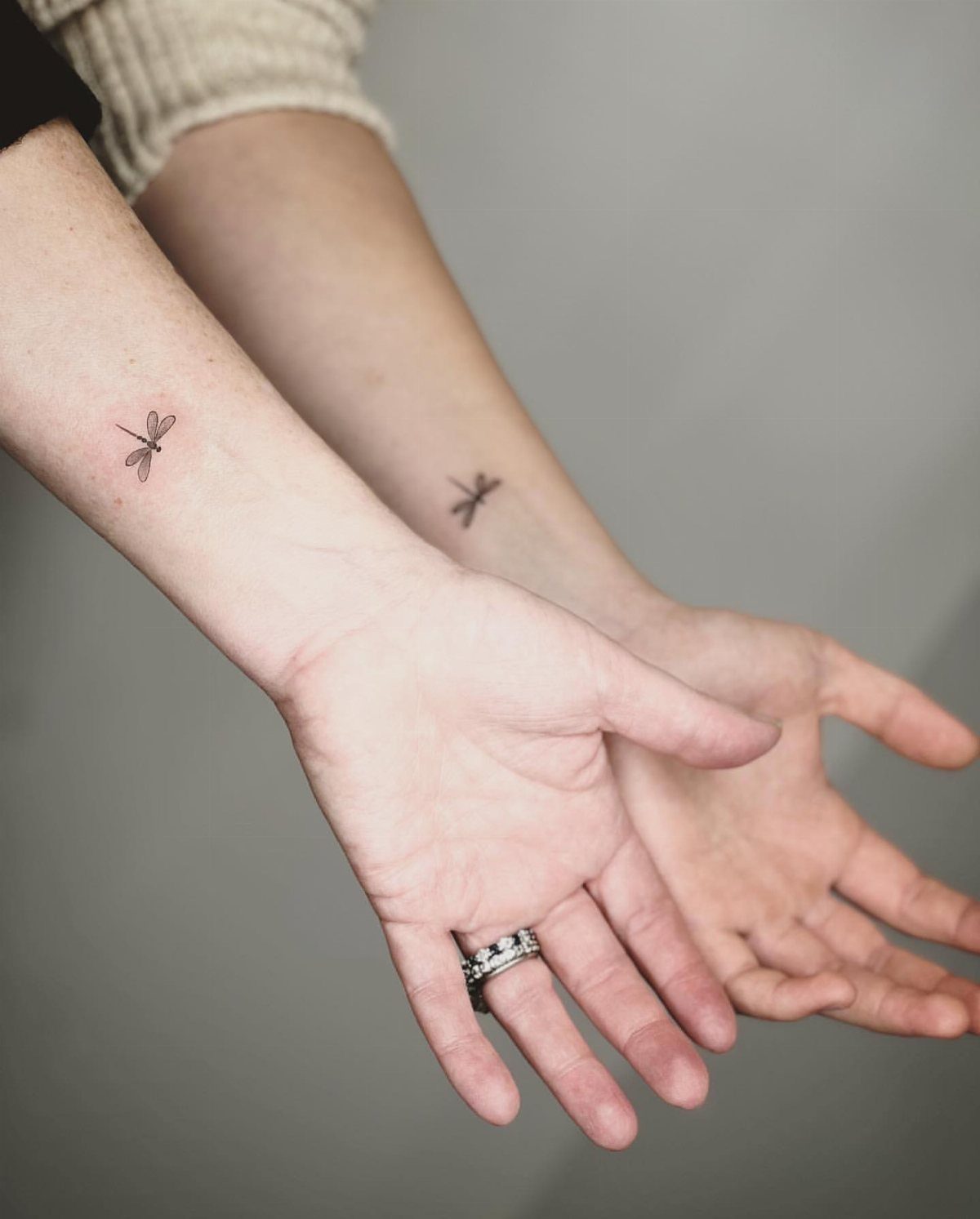 Two inner wrists featuring matching small dragonfly tattoos against a grey background