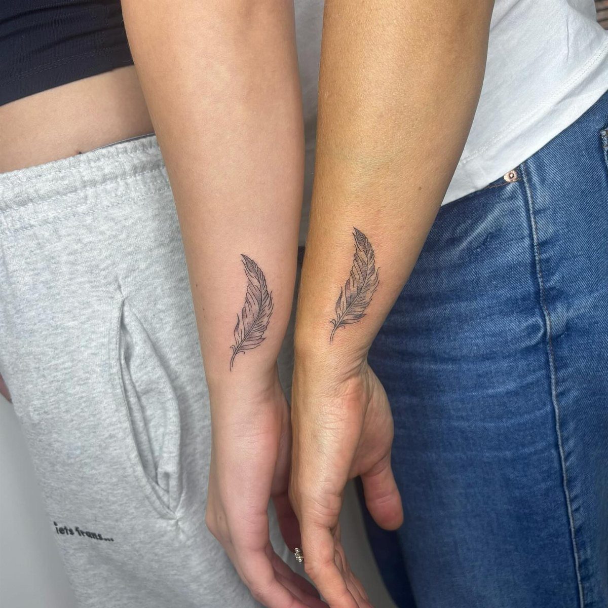 Matching black feather tattoos on the wrists of two people standing together.