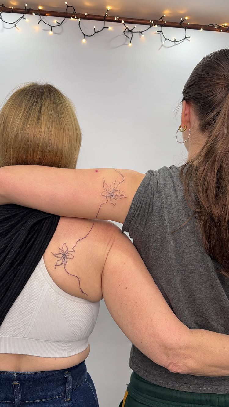 Two women with matching fine-line flower tattoos on their arm and shoulder.