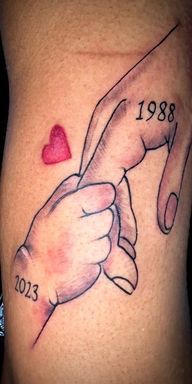 Tattoo of an adult hand holding a baby's hand with a red heart.