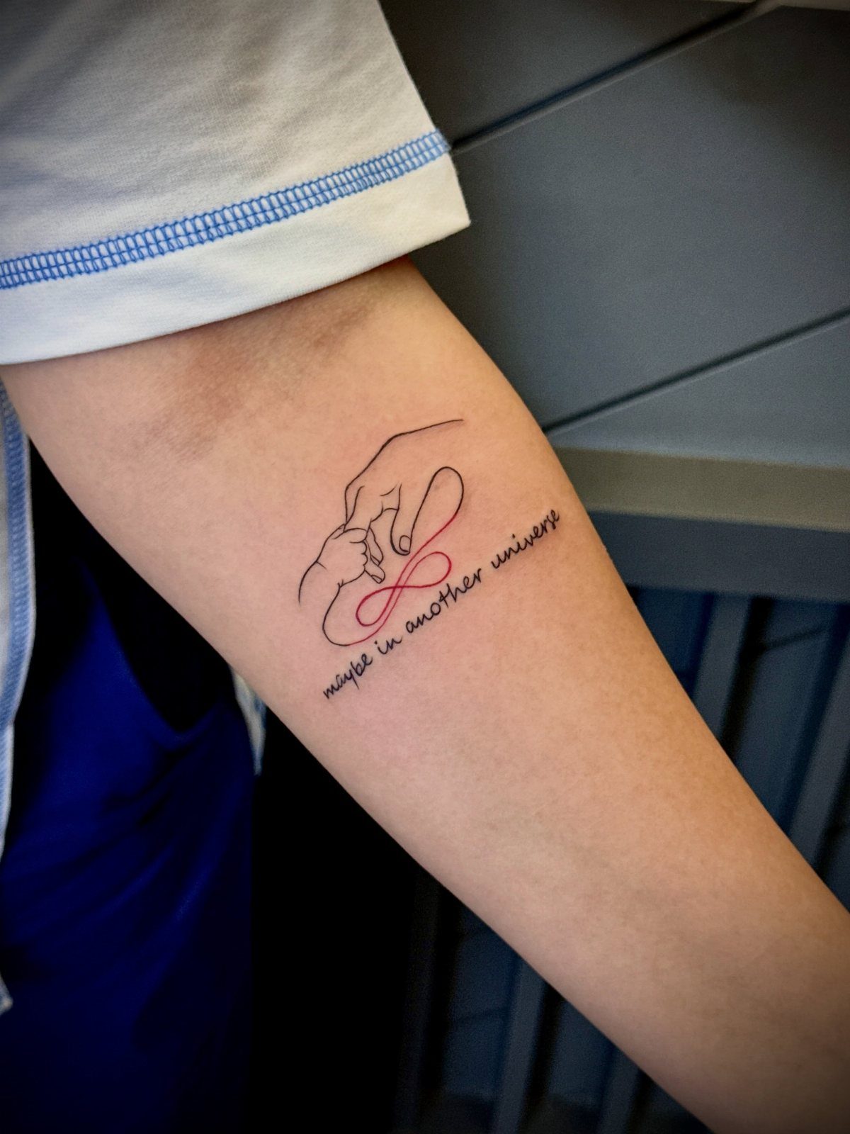 Tattoo on a forearm showing an adult and child holding hands with red infinity symbol.