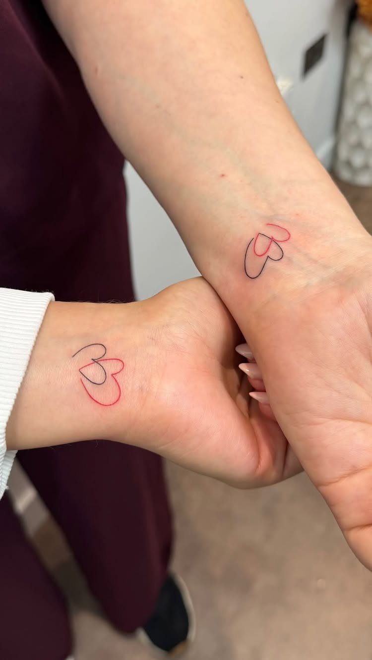 Two wrists with matching tattoos of overlapping black and red heart outlines.