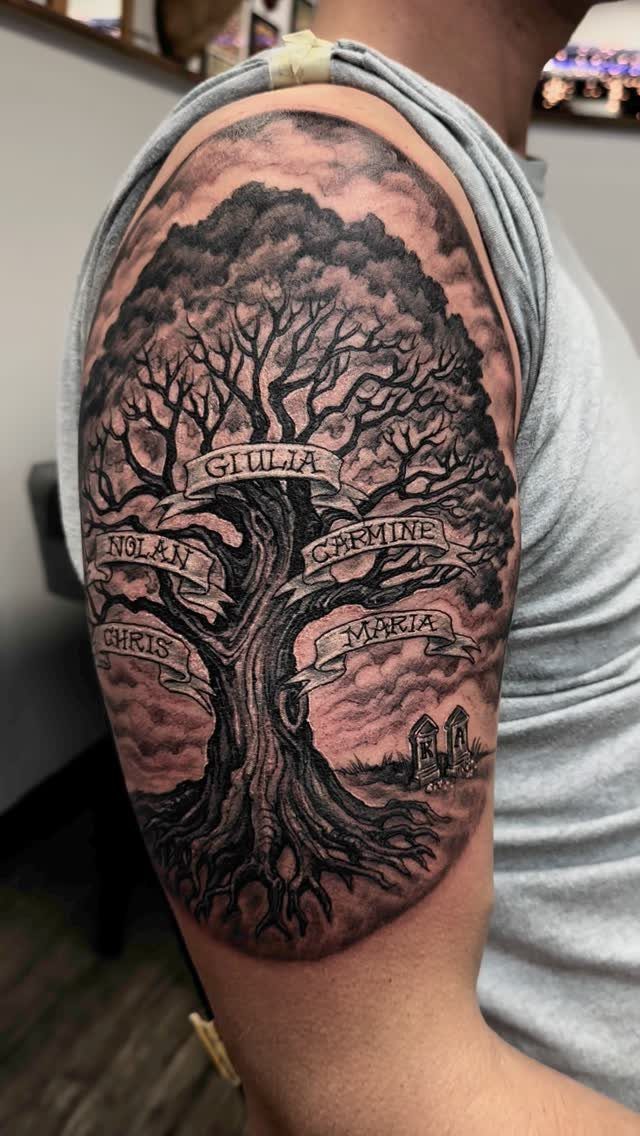 Family tree tattoo on an upper arm with names on banners and headstones.