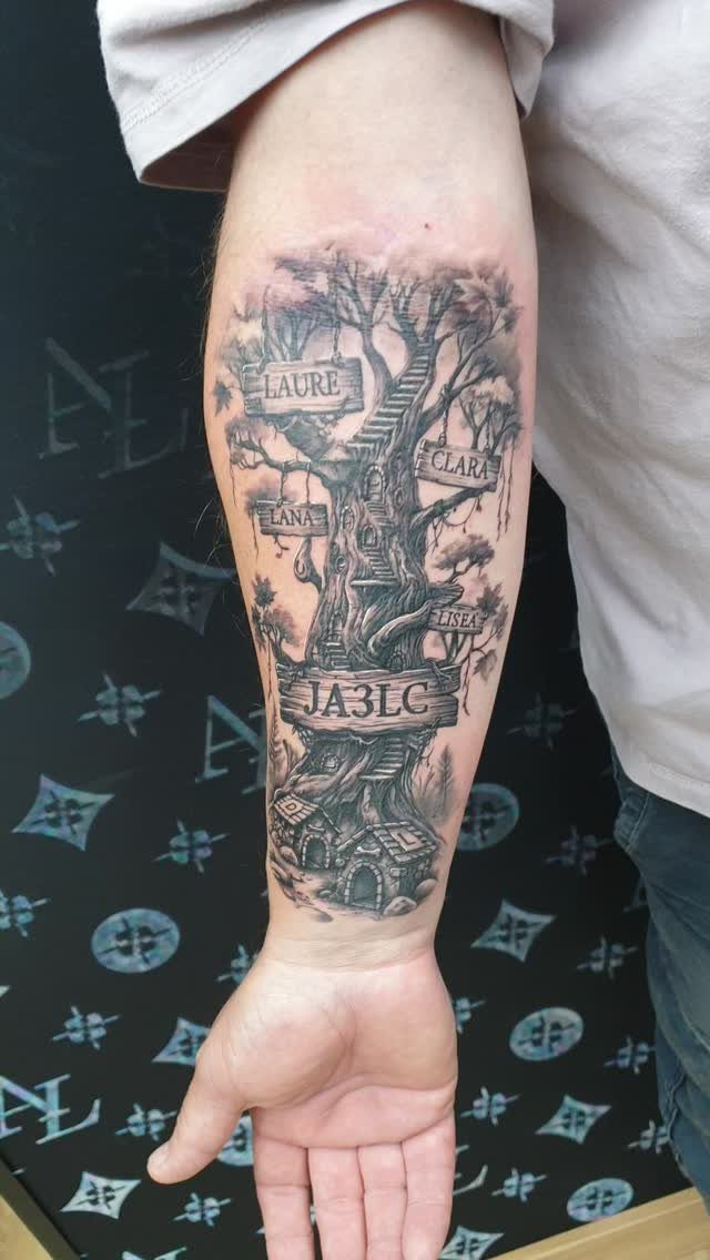 Detailed black and grey family tree tattoo on a forearm with names on banners.