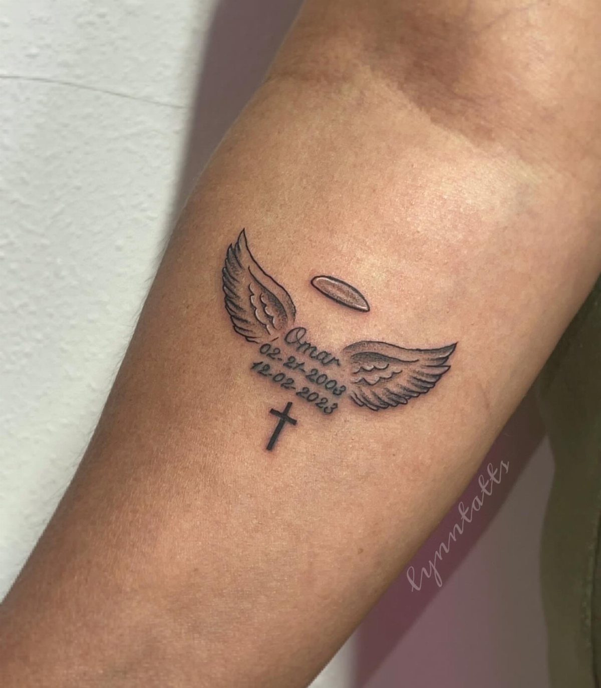 Memorial tattoo on forearm with angel wings, halo, cross, and name Omar.