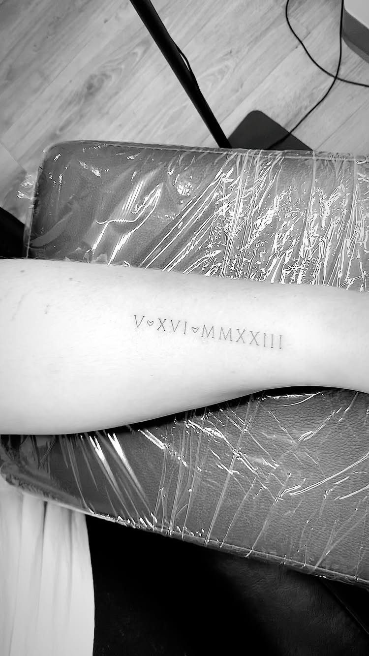 Roman numeral birthdate tattoo with small hearts on a person's forearm