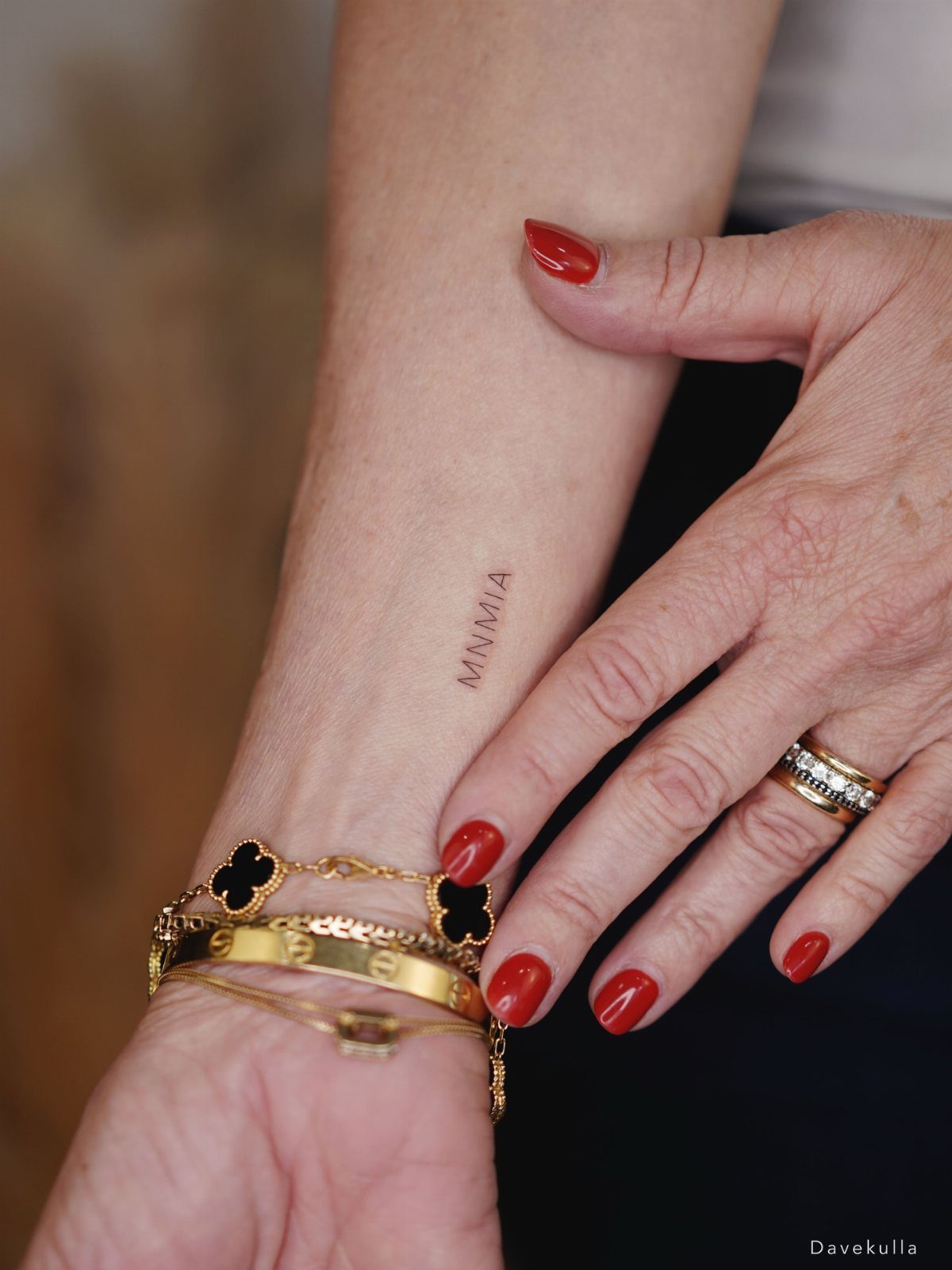 Small black ink initials tattoo on a wrist with gold jewelry and red nails