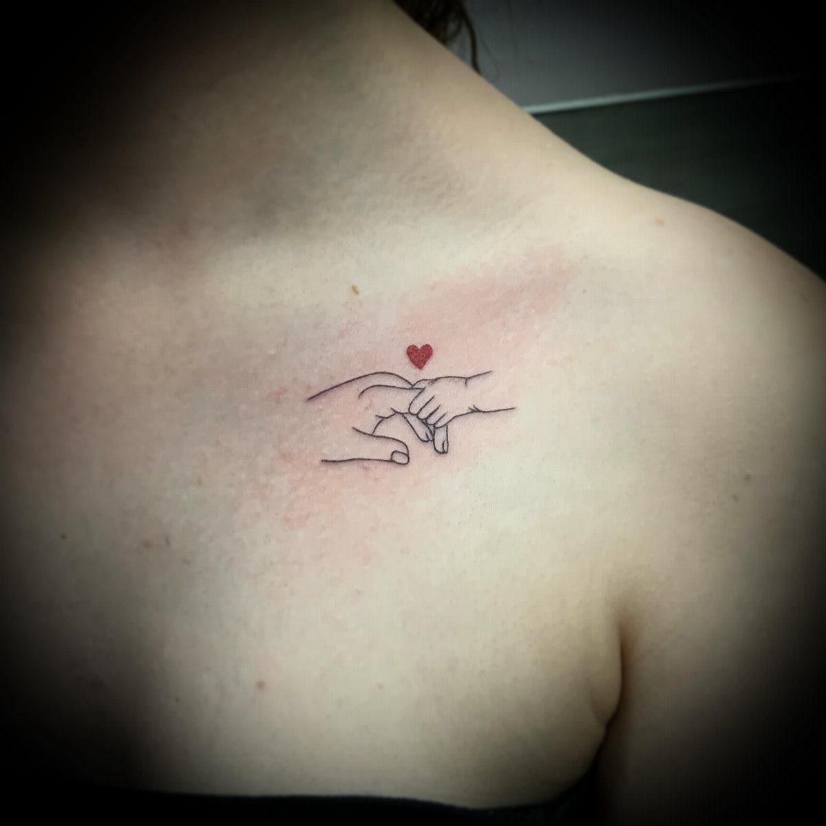 Minimalist line-art tattoo of two pinky-swearing hands with a small red heart.