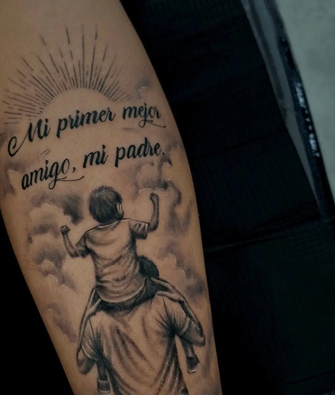 Tattoo of a child on a father's shoulders with Spanish text and sunburst.