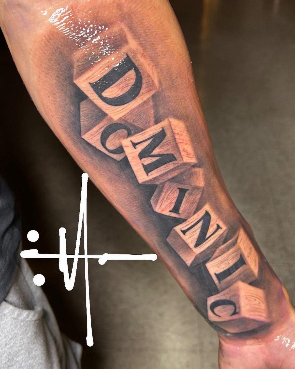 Tattoo of the name Dominic spelled out using 3D baby blocks on a forearm.