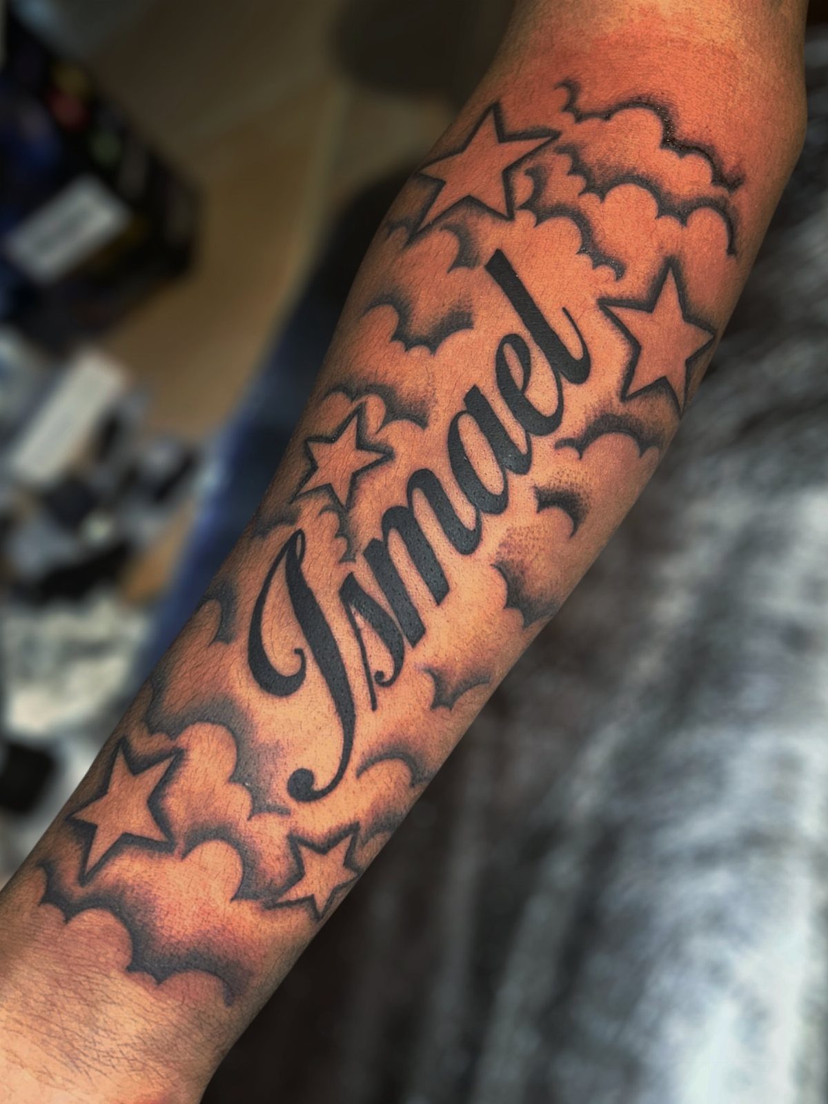 Cursive name Ismael tattoo on a forearm surrounded by stars and clouds