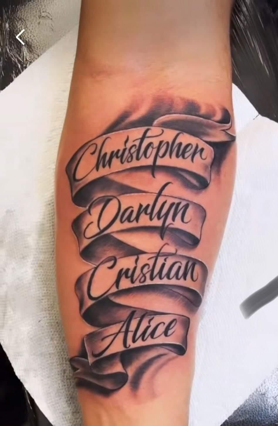 Tattoo on a forearm showing four names written inside a flowing ribbon banner.