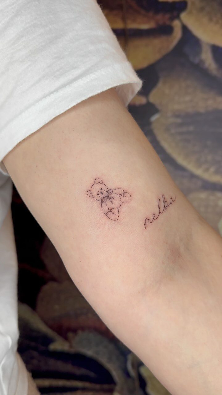 Teddy bear and cursive name melba tattooed on a person's inner forearm.