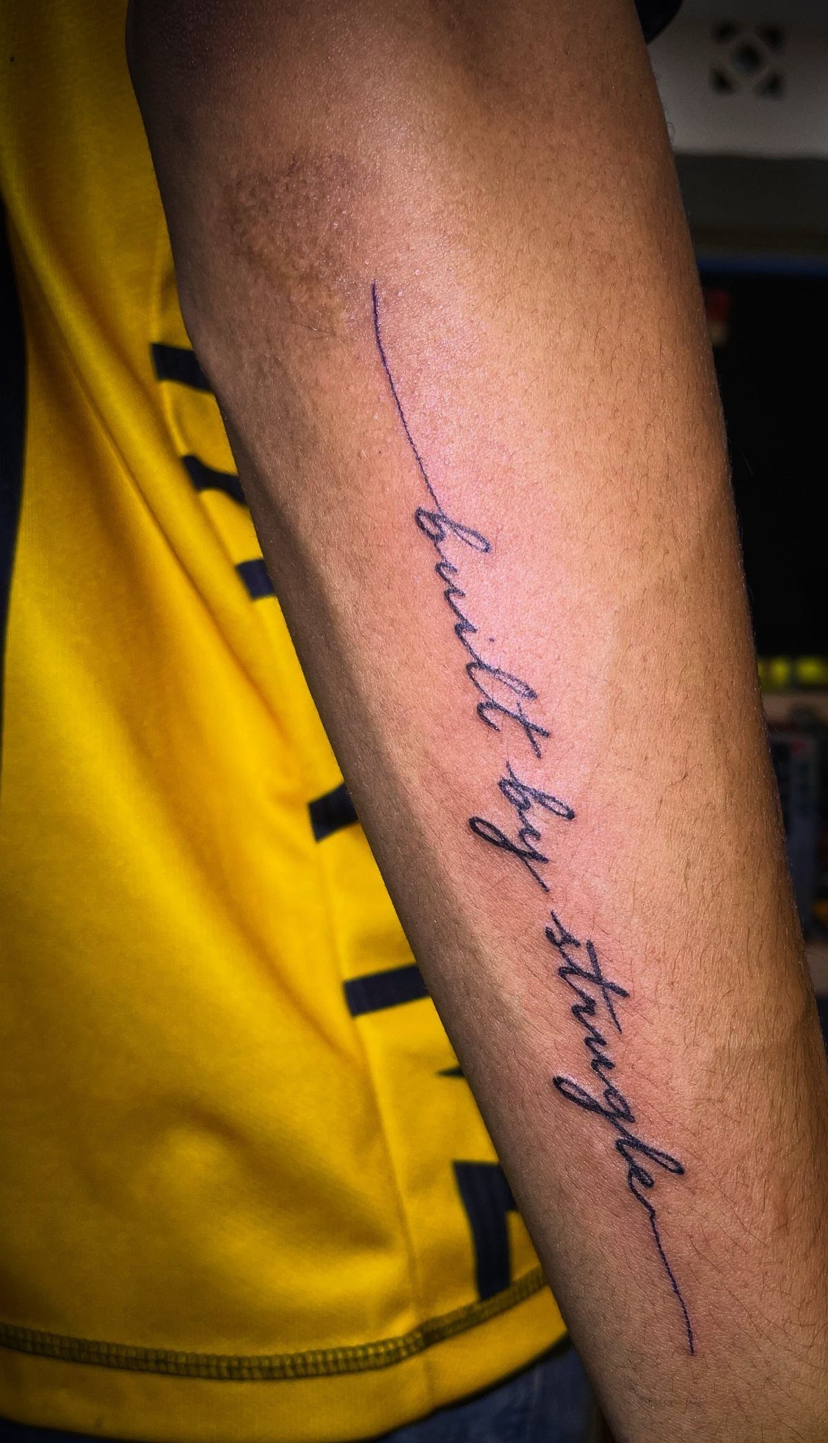 Cursive tattoo on a forearm reading built by struggle in thin black ink