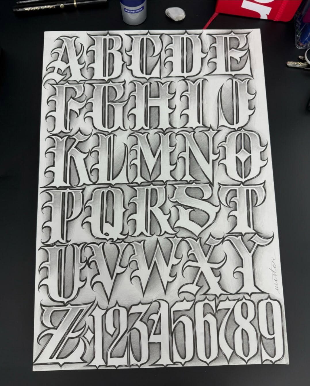 Gothic tattoo lettering alphabet and numbers drawn with grey shading on white paper.