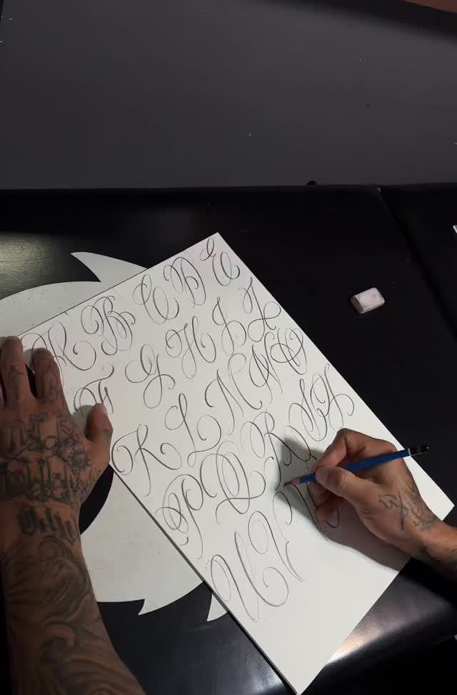 Hand-drawn script alphabet calligraphy on white paper with tattooed hands holding a pen