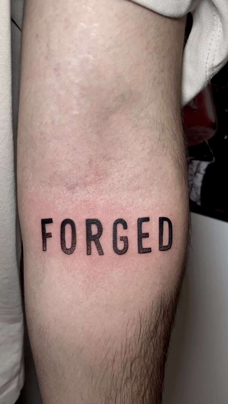 Black ink tattoo of the word FORGED in bold lettering on a forearm