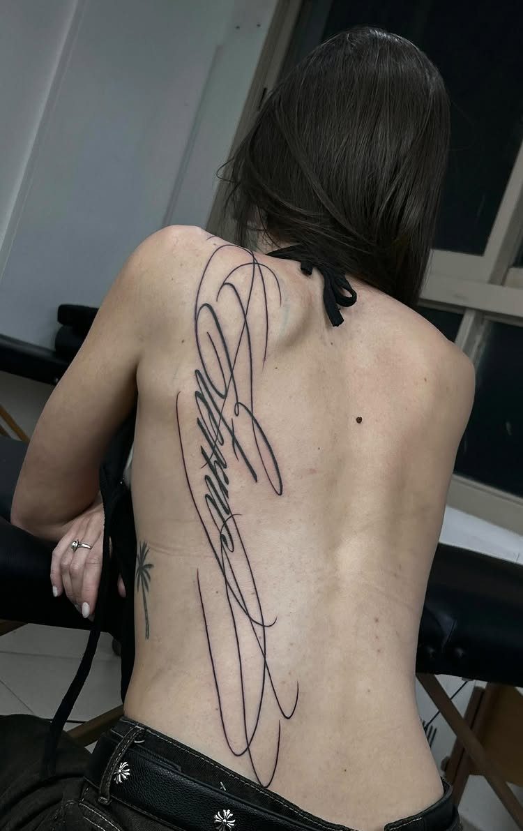Woman's back with a large vertical flowing cursive script tattoo in black ink.