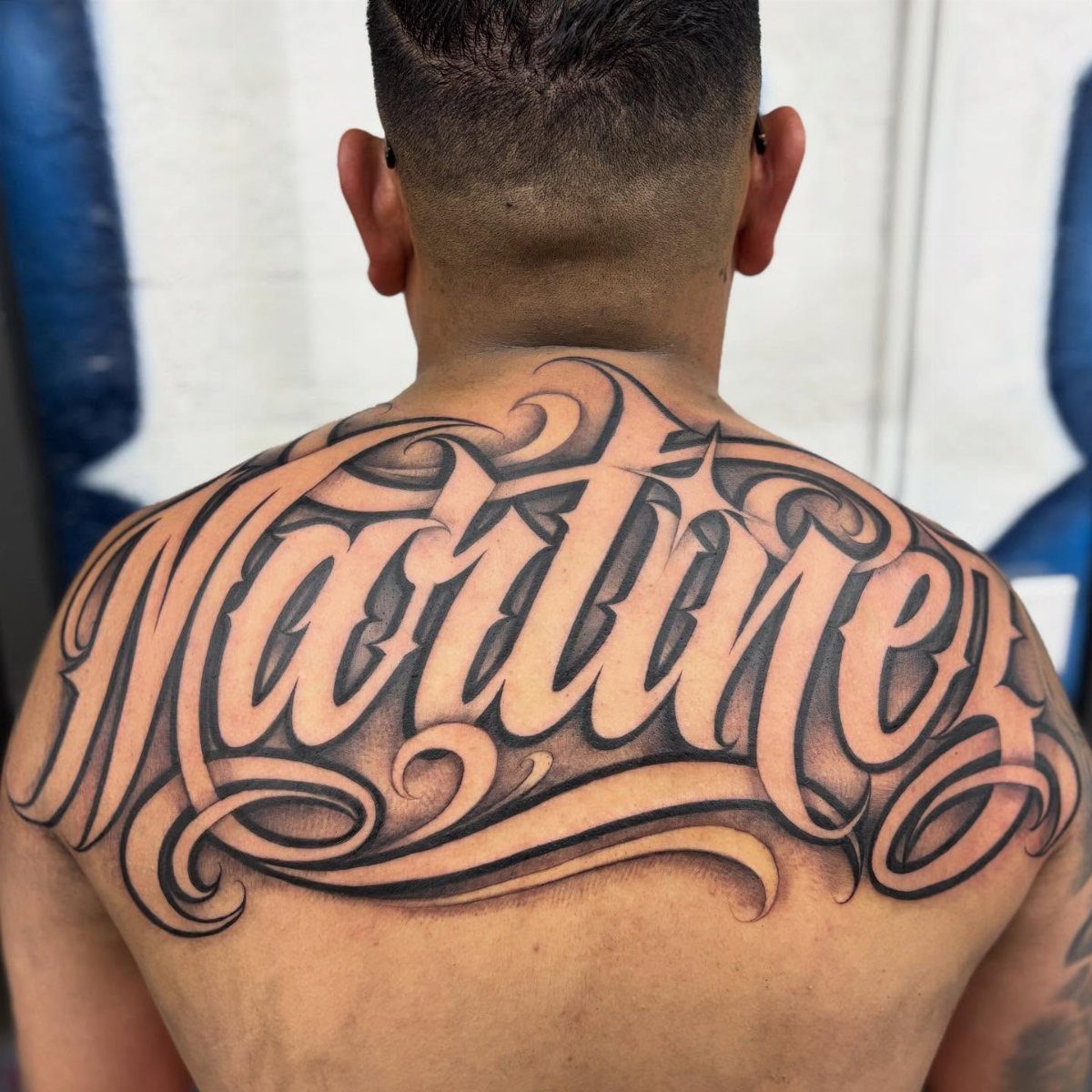 Large stylized Martinez name tattoo across the upper back of a man.