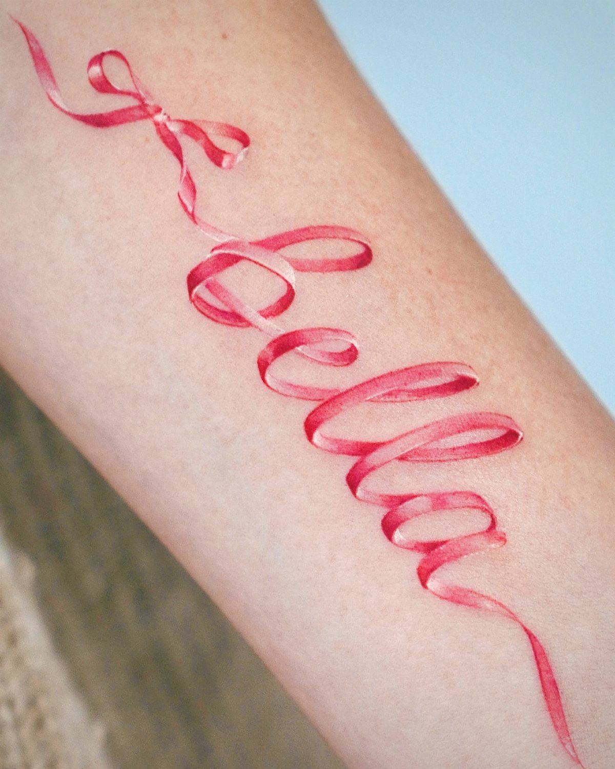 Red ribbon tattoo on an arm spelling the name Bella with a bow.