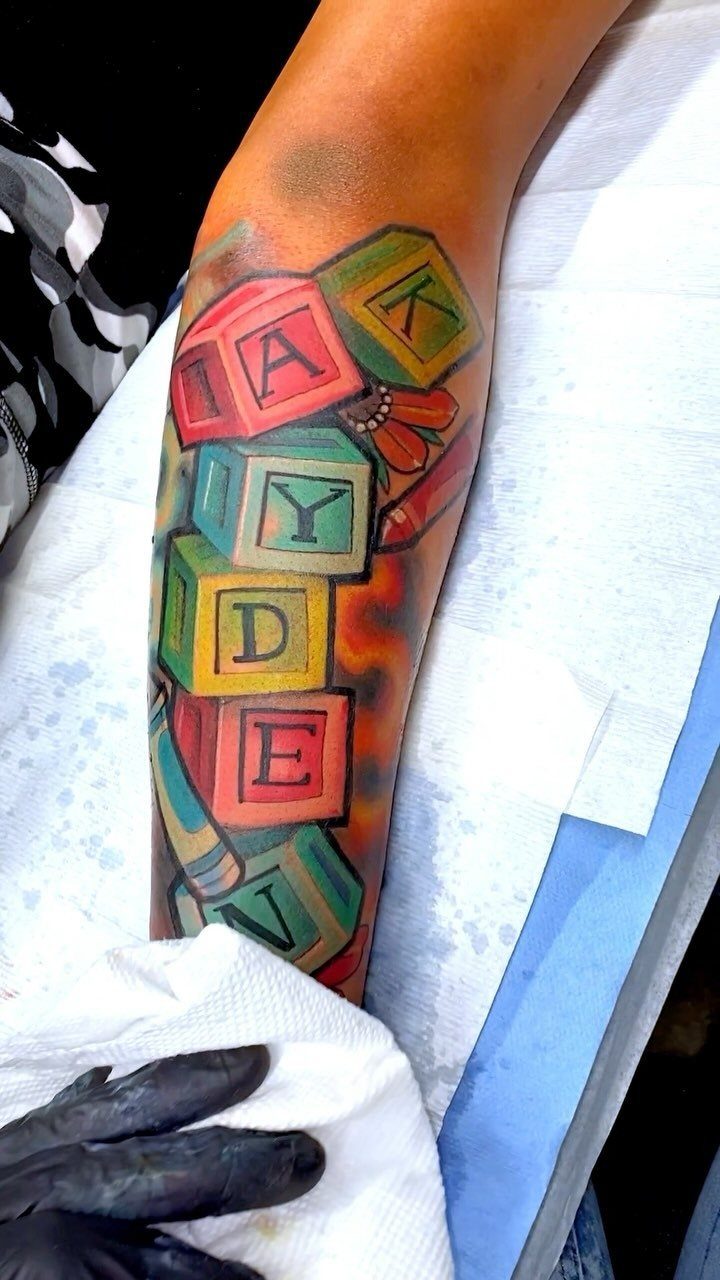 Colorful tattoo of alphabet blocks spelling KAYDEN on a person's forearm