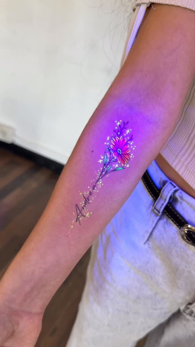 Colorful flower tattoo with a cursive name integrated into the stem on a forearm