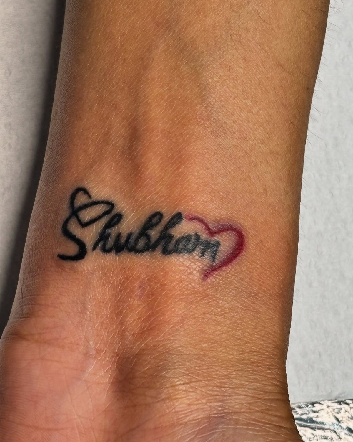 Cursive tattoo of the name Shubham with a red heart on a wrist