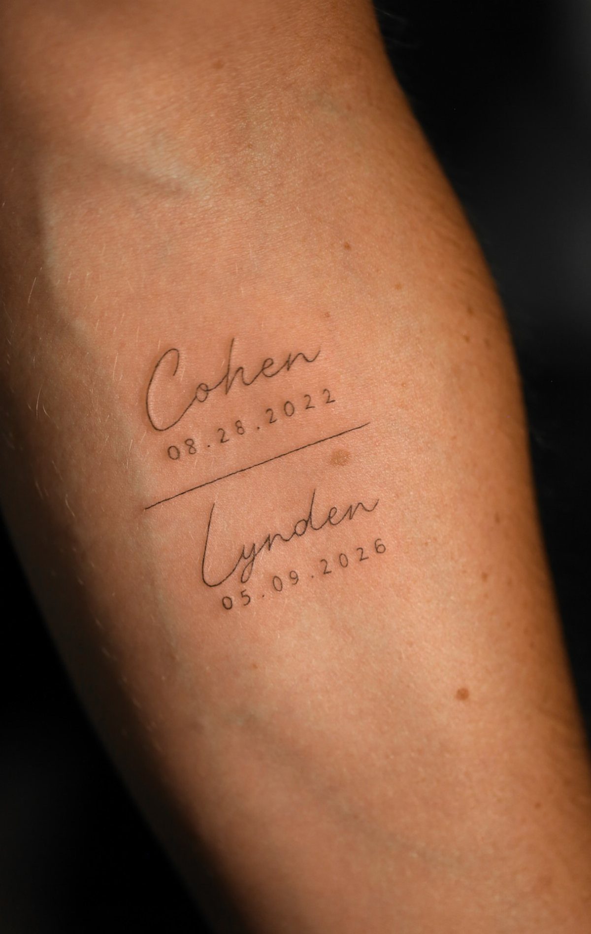 Cursive name tattoos of Cohen and Lynden with dates on a forearm