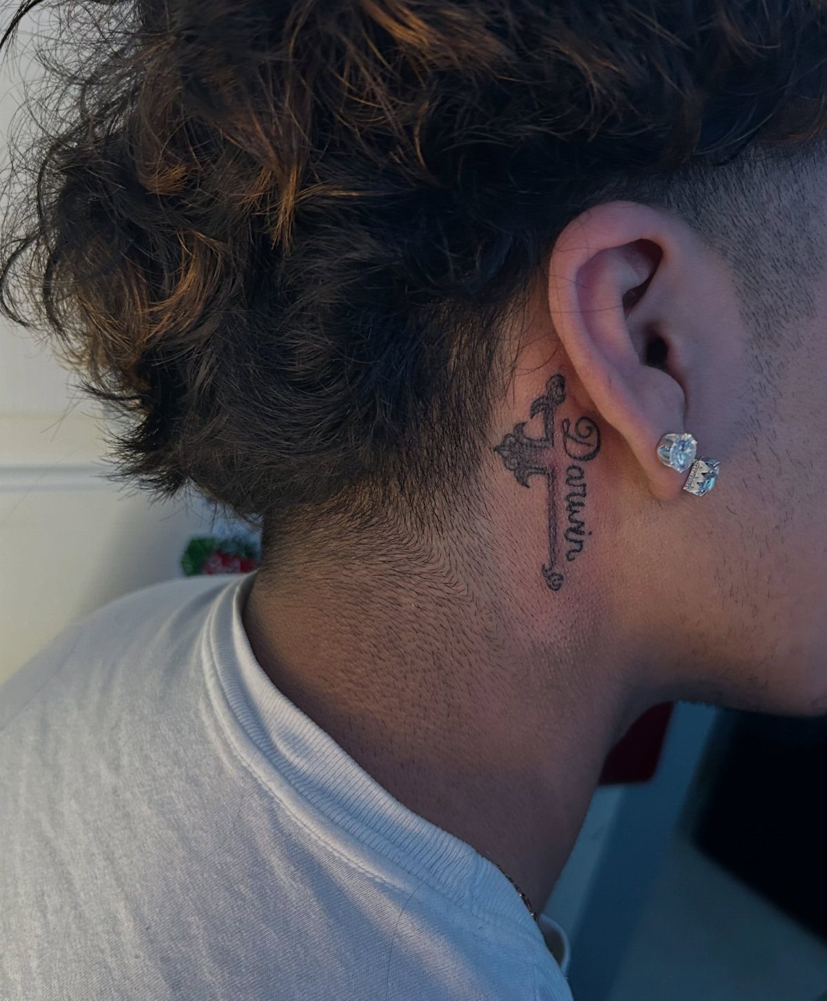 Tattoo of a cross and the name Darwin located behind a person's ear.