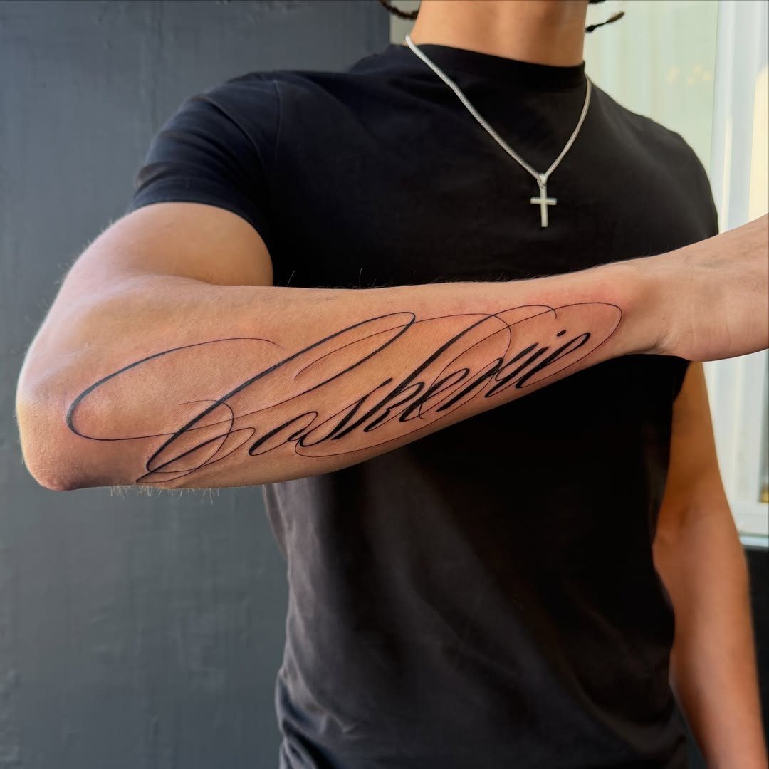Cursive name tattoo on a person's forearm with a black t-shirt background
