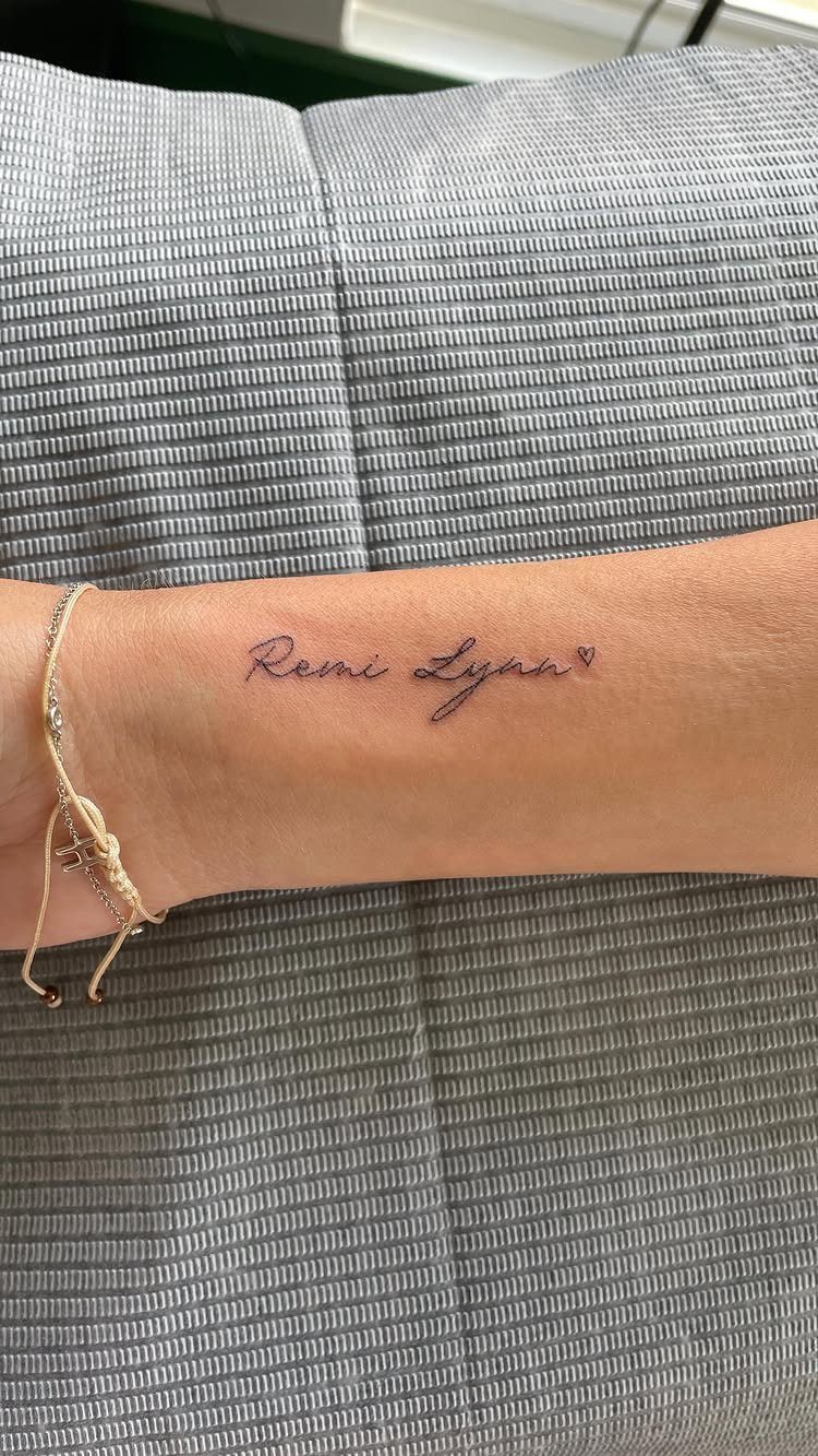 Cursive name tattoo on a wrist with a small heart and gold bracelet.
