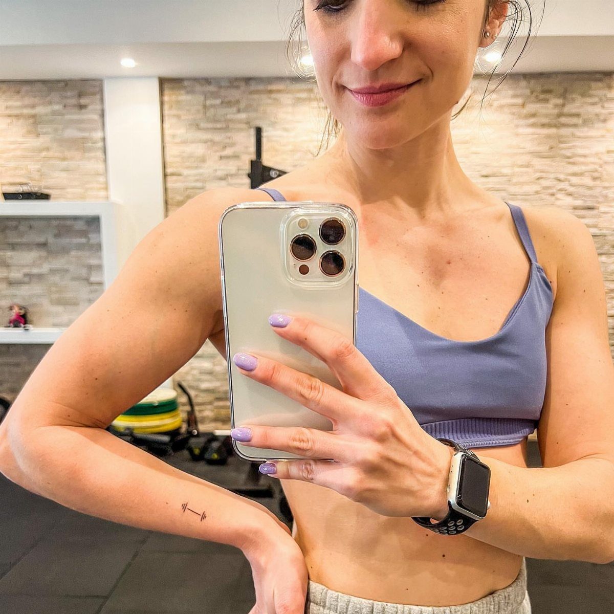 Woman in gym attire with a small barbell tattoo on her inner forearm.
