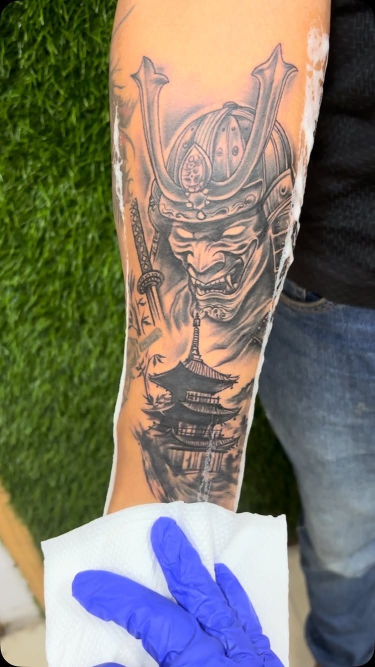 Samurai mask tattoo with a pagoda and sword on a person's forearm