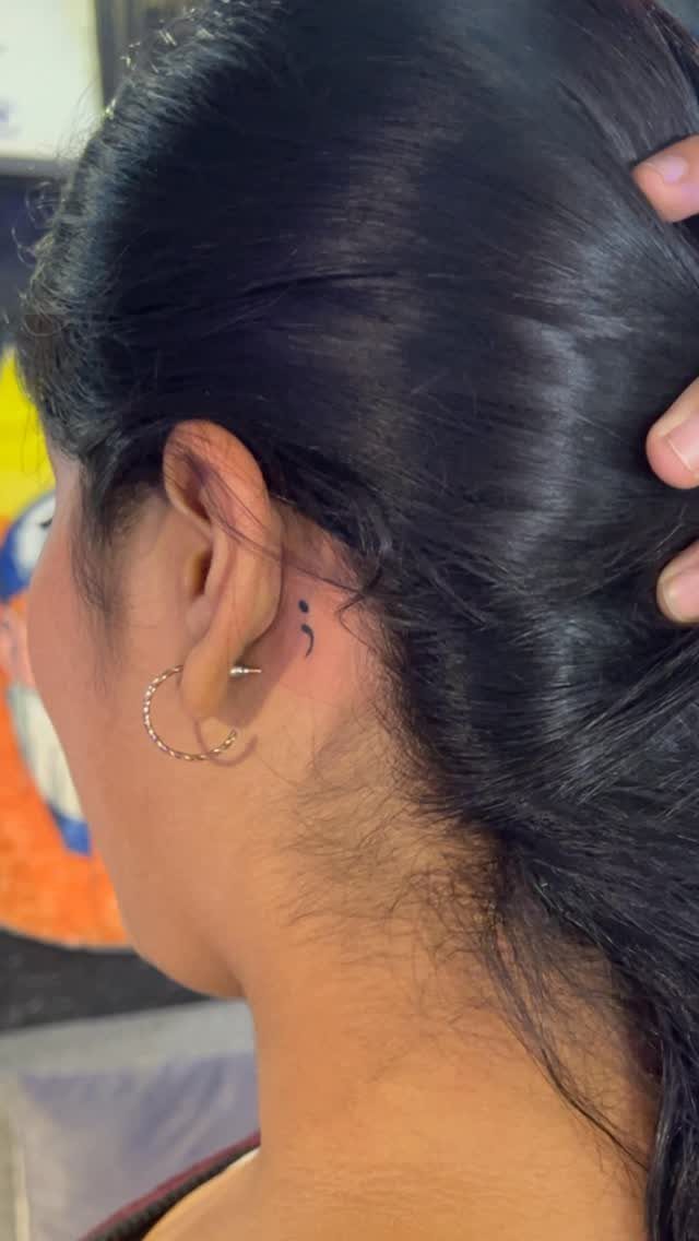 Semicolon tattoo located behind the ear of a person with black hair.