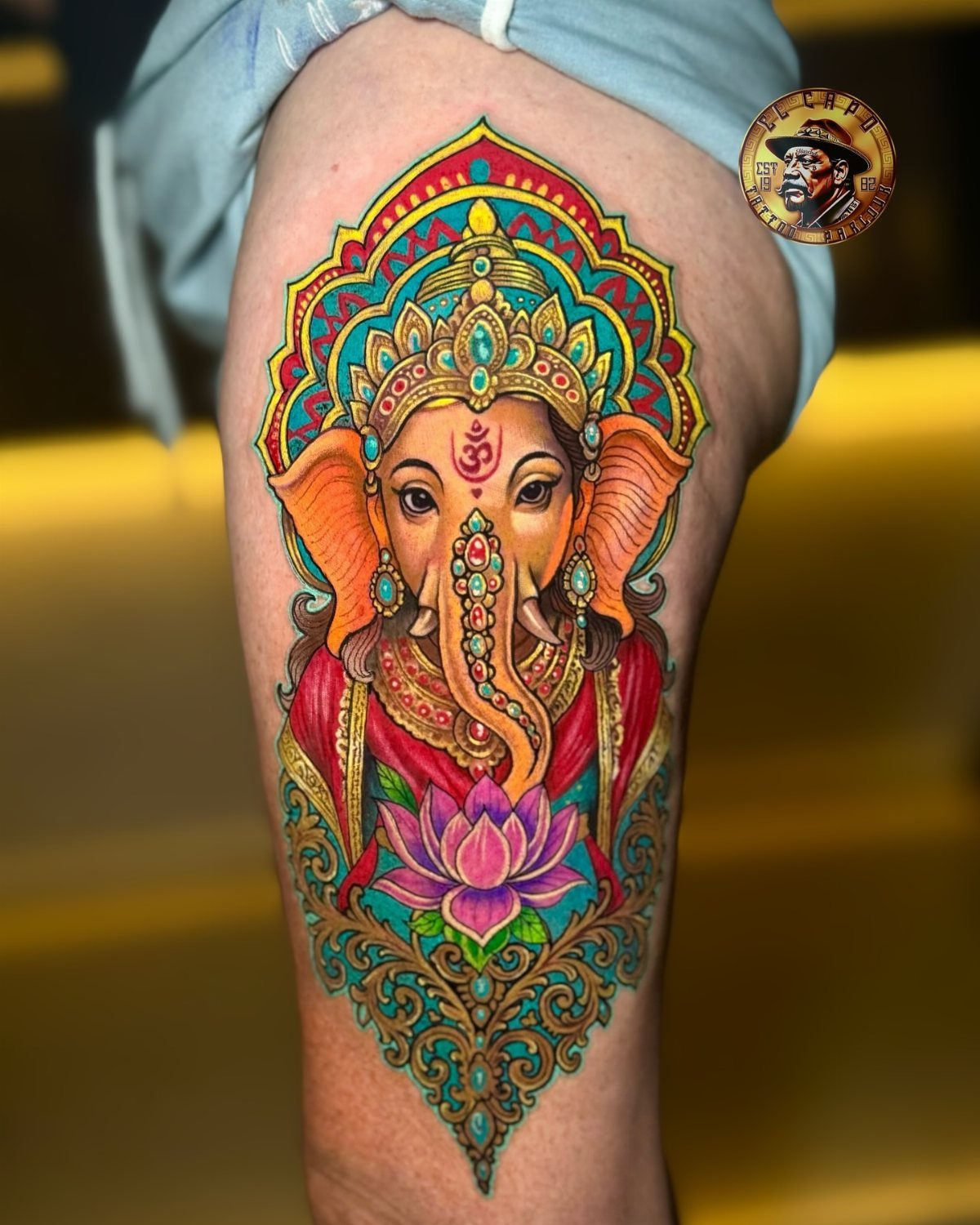 Colorful Ganesha tattoo on a leg featuring an ornate crown and pink lotus.