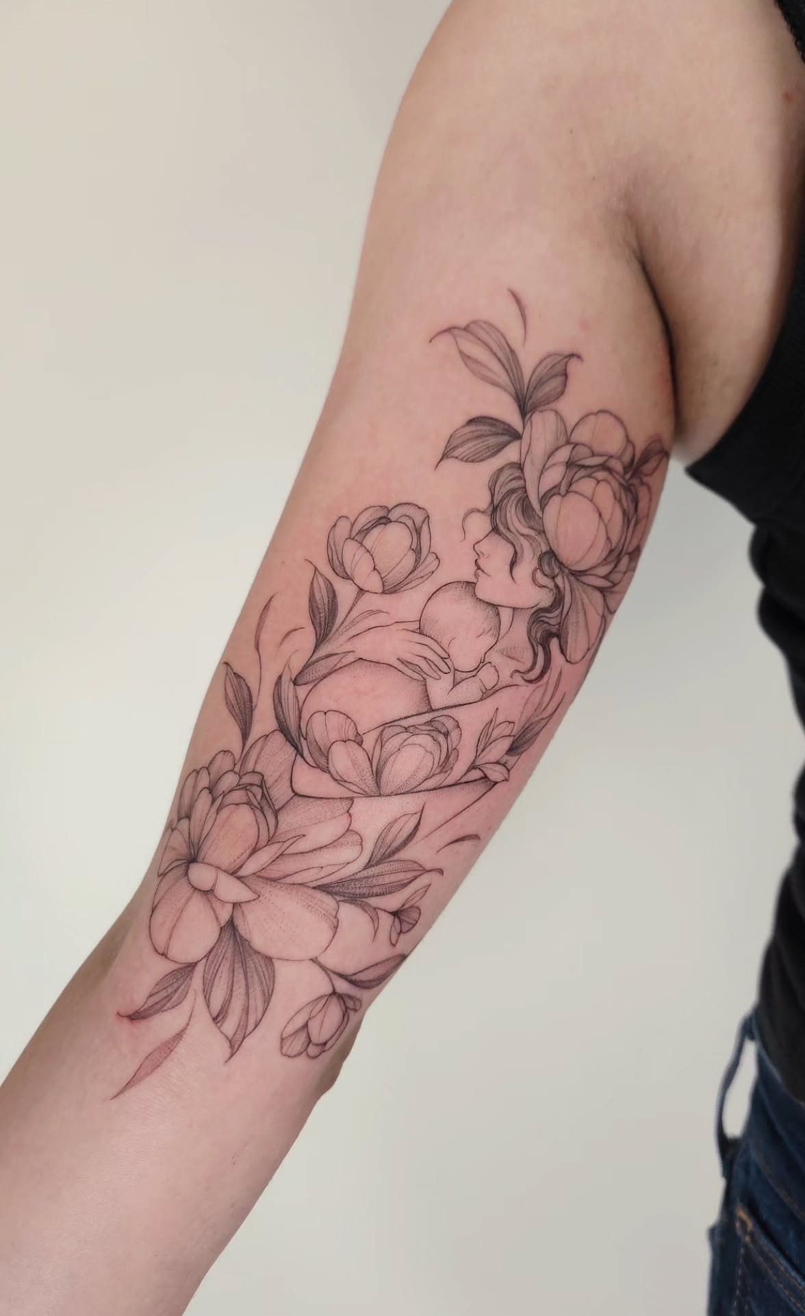 Forearm tattoo of a woman holding a baby surrounded by peony flowers.
