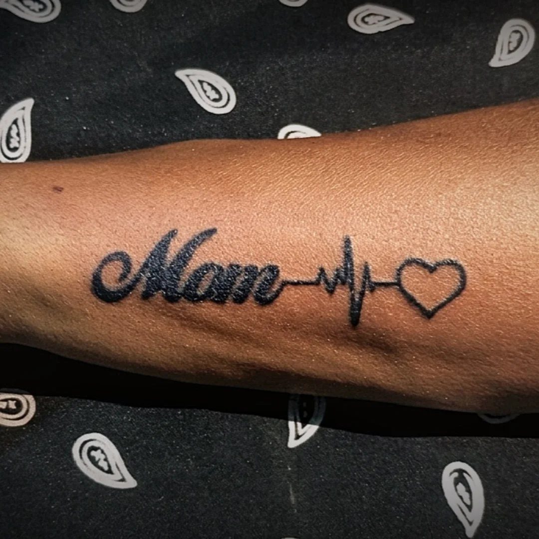 Black ink tattoo on a forearm reading Mom with a heartbeat and heart.