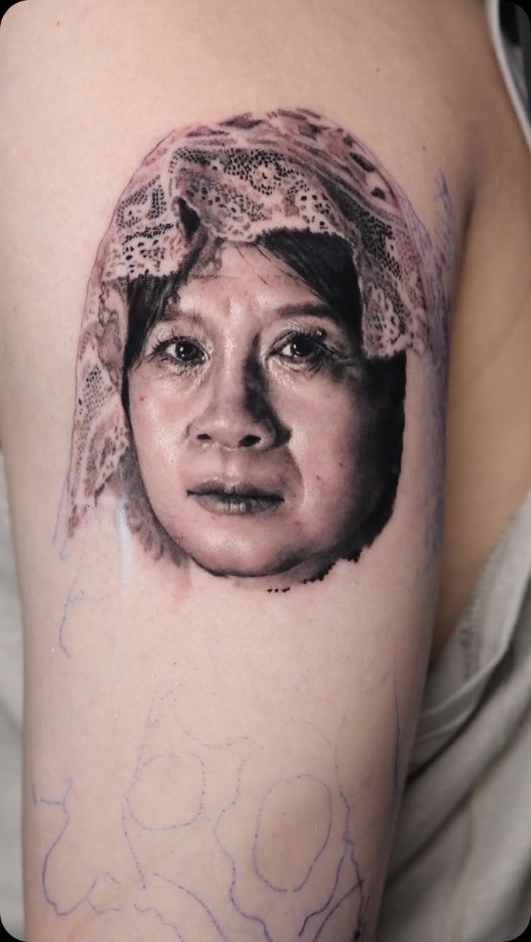 Realistic portrait tattoo of an older woman with a lace head covering on an arm.