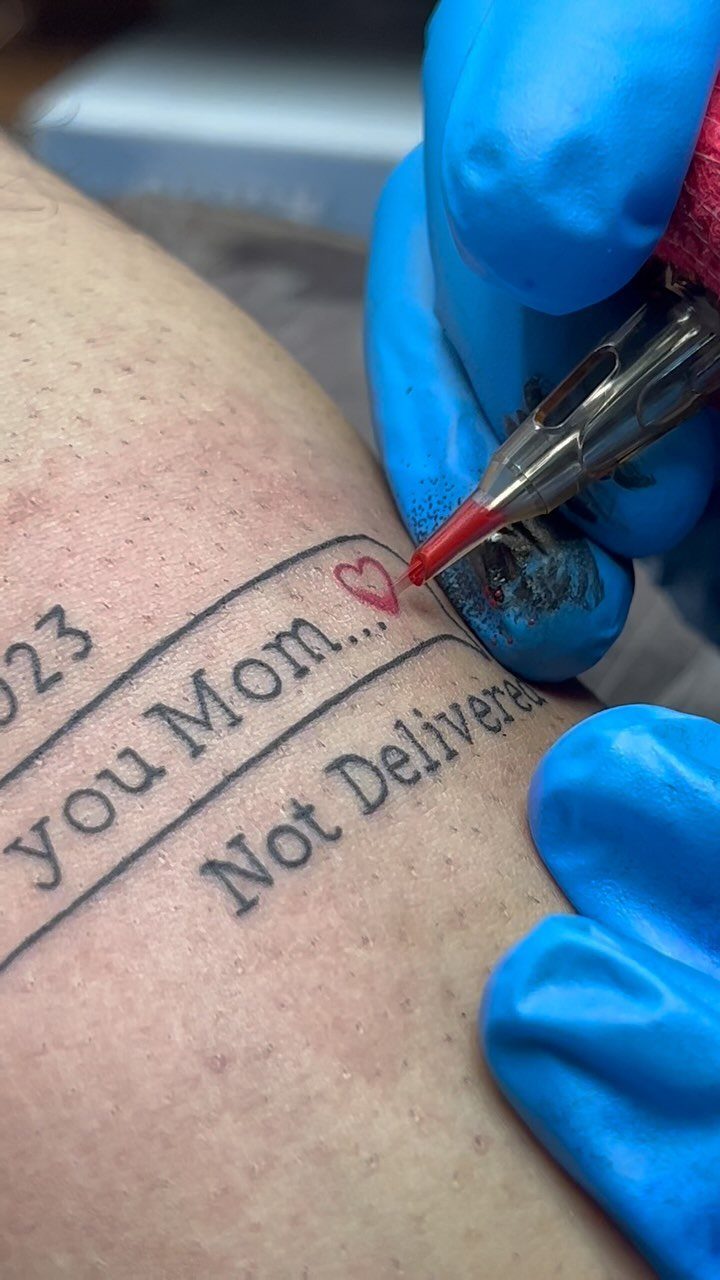 Tattoo artist in blue gloves tattooing the words you Mom with a red heart