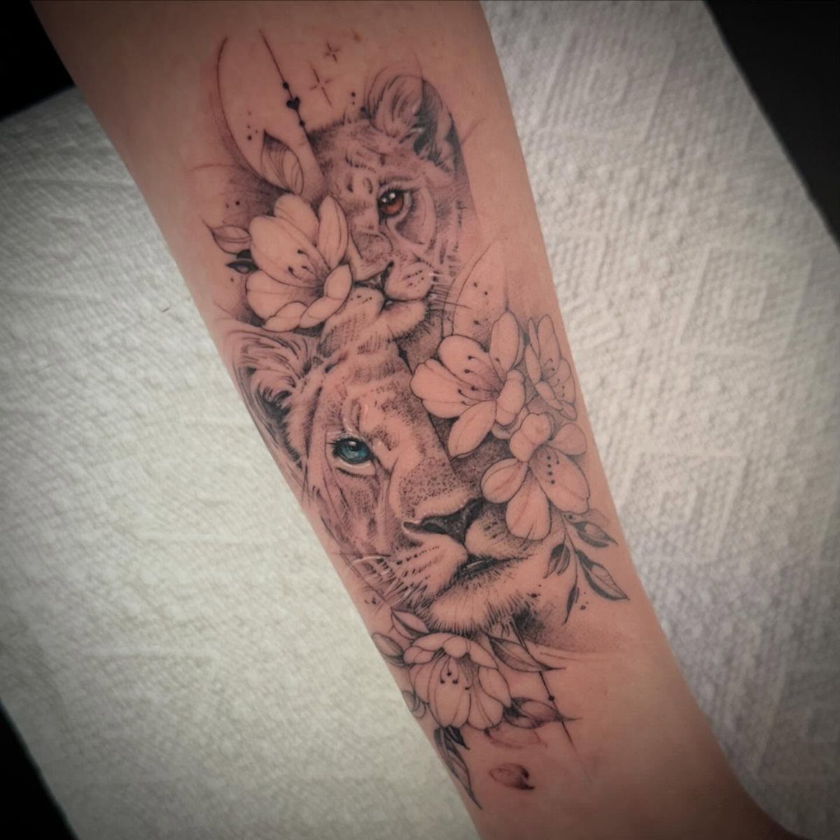 Lion tattoo on a forearm featuring two lion faces surrounded by delicate flowers.