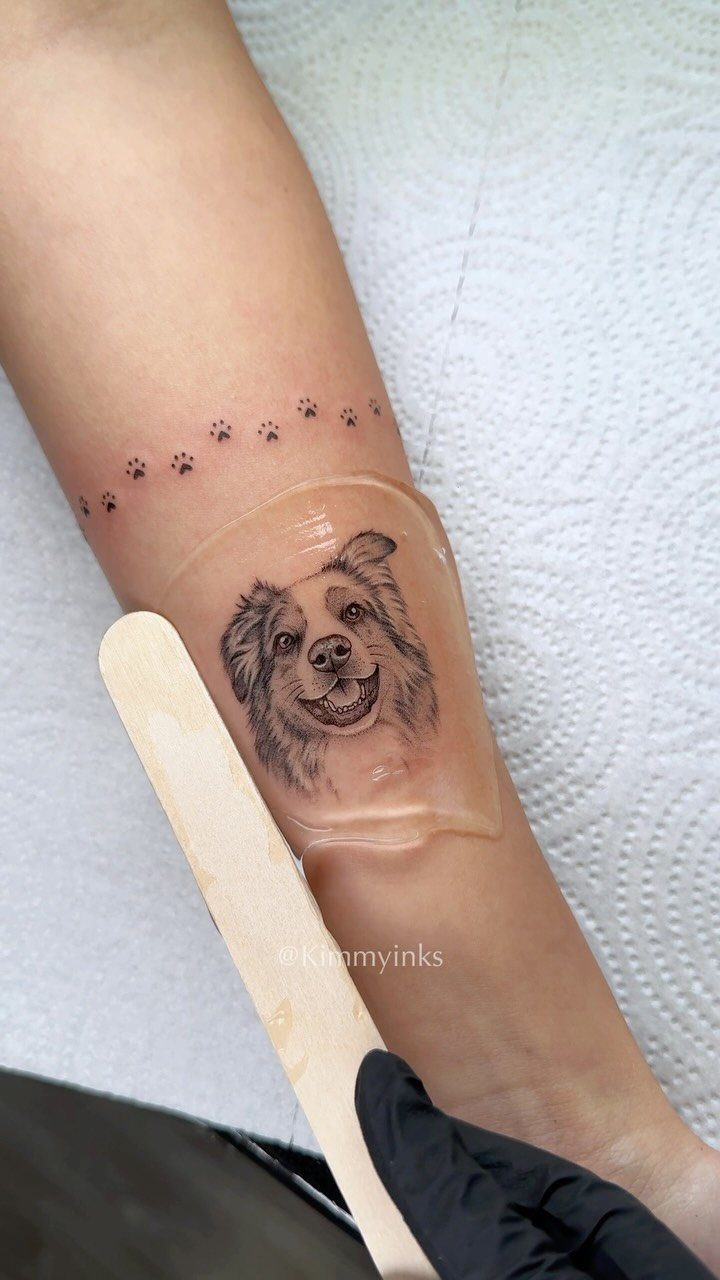 Realistic dog portrait tattoo on a forearm with a row of small paw prints.