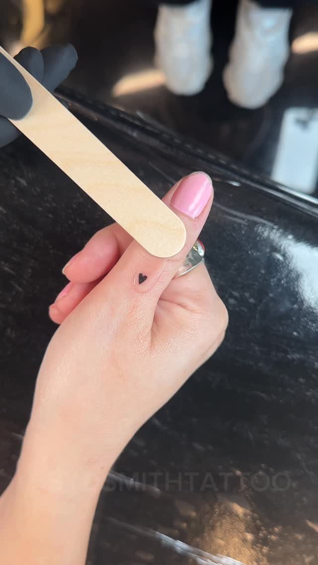 Small black heart tattoo on the side of a finger with pink nails