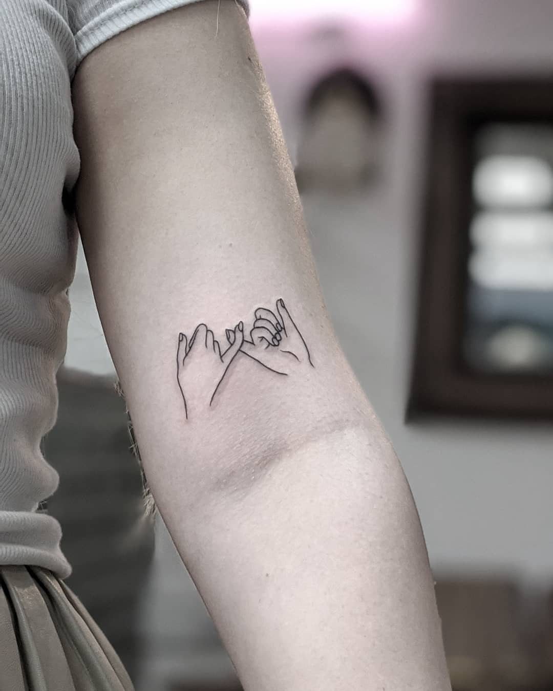 Minimalist black ink tattoo of two hands interlocking pinky fingers on a forearm.