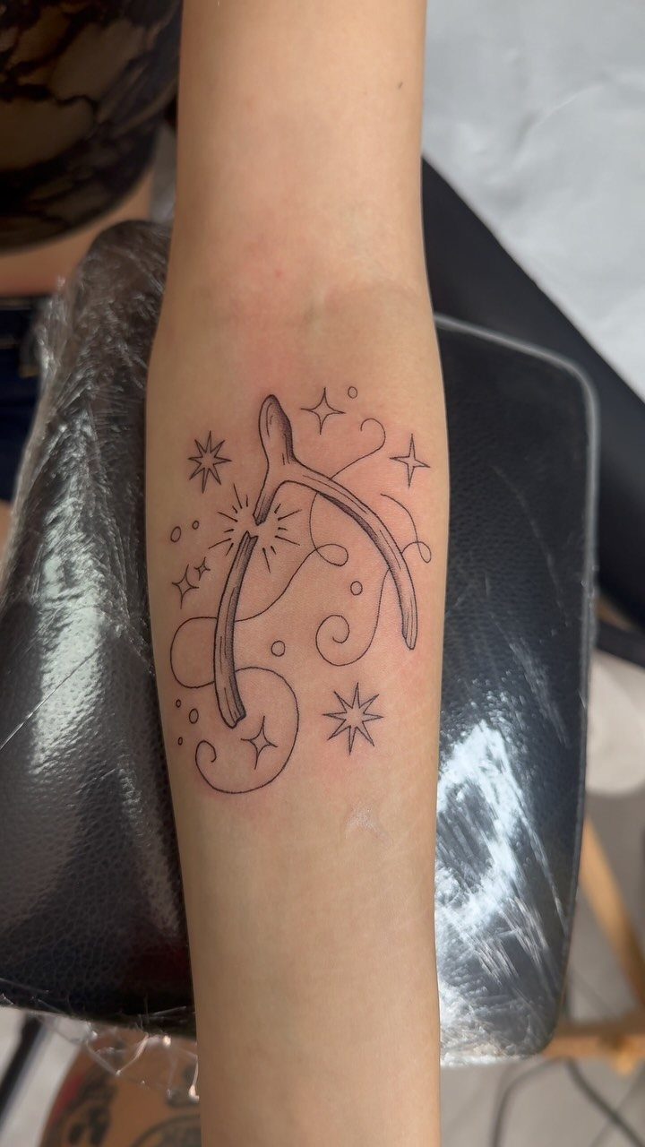 Wishbone tattoo on a forearm with decorative stars and swirls in black ink.