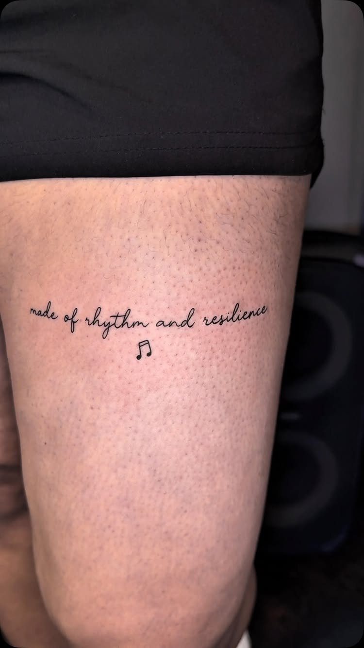 Cursive tattoo on a thigh reading made of rhythm and resilience with musical note