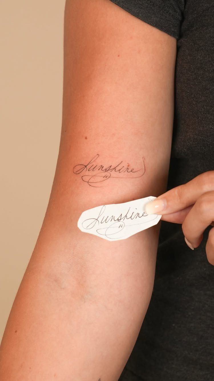 Script tattoo reading Sunshine on a person's inner forearm with a paper template.