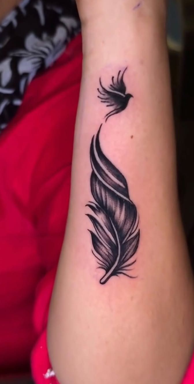 Black ink tattoo of a detailed feather and a small bird on a forearm.